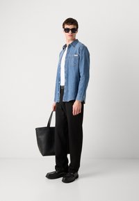 Blauwe denim overhemd, witte t-shirt, zwarte broek en zwarte schoenen. Houdt een zwarte tote bag vast. Zonnebril maakt de look compleet.