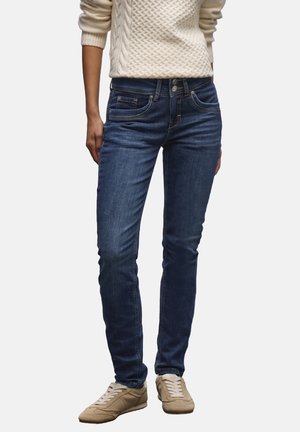Personne portant un jean slim bleu foncé, des baskets beiges et un pull blanc texturé avec les manches retroussées.