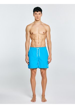 Fit młody mężczyzna stoi boso, ubrany w jasnoniebieskie szorty kąpielowe z białym troczkiem na prostym jasnym tle.