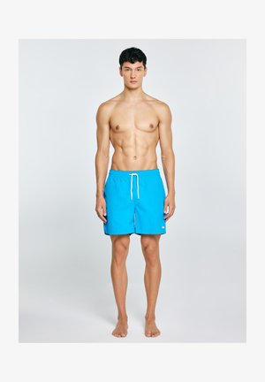 Fit młody mężczyzna stoi boso, ubrany w jasnoniebieskie szorty kąpielowe z białym troczkiem na prostym jasnym tle.