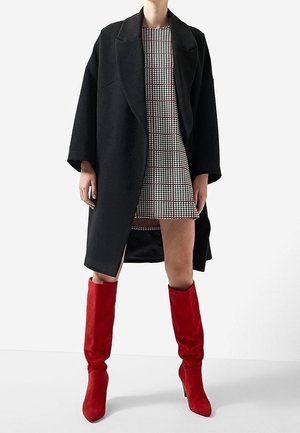 Osoba ubrana w czarny, za duży płaszcz na mini sukience w jodełkę, do tego czerwone kozaki na obcasie sięgające do kolan, na jednolitym tle.