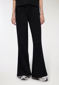 Pantaloni da tuta neri a gamba larga con cintura a cordino, tasche e una texture morbida; abbinati a sneaker argentate riflettenti.