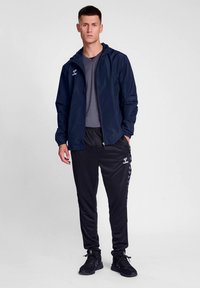 Tamsiai mėlynas vėjo nepraleidžiantis striukė su kapišonu, pilka marškinėliai ir juodos sportinės kelnės su šonine juostele, baigtos juodais sportbačiais.