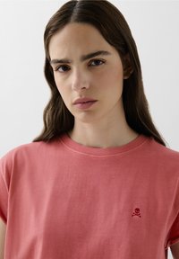 T-shirt in cotone rosa con scollatura rotonda e maniche corte, caratterizzata da un piccolo teschio e ossa incrociate ricamati sul petto.