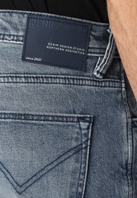 Jeans iz denima svetle modre barve, ki imajo usnjen trak z oznako "DENIM TOM TAILOR" na zadnjem pasu. Vidna sta obrabljena tekstura in podrobnosti šivanja.