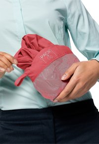 Röd packbar väska med nätbotten och elastiskt topp, med "PACK + GO"-branding, hålls i en hand. Väskan är lätt och kompakt.