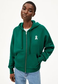 Grüne Zip-Up-Trainingsjacke aus weichem Stoff mit einer Fronttasche, Kordelzügen und einem weißen Ribbons-Logo auf der Brust. Kombiniert mit blauen Jeans.