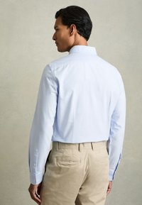 Muškarac u svijetloplavoj košulji dugih rukava i bež hlačama, stoji bočno okrenut lijevo na neutralnoj pozadini.