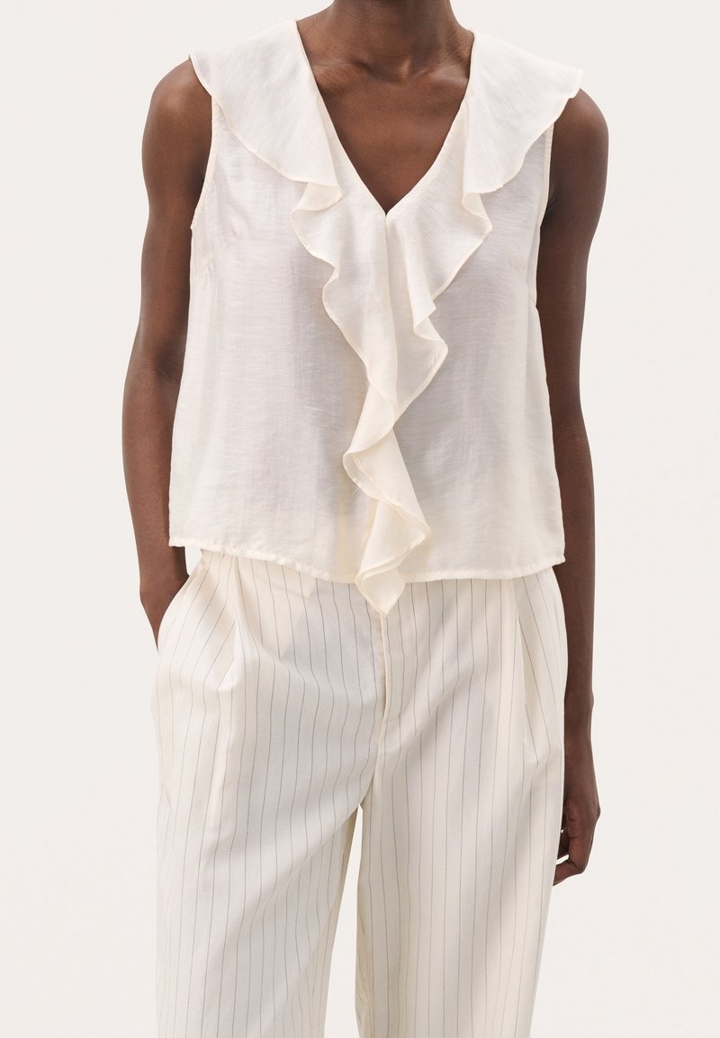 Femme portant un chemisier blanc sans manches avec un détail de volants à l'avant et un pantalon blanc taille haute à fines rayures verticales, les mains dans les poches.