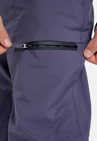Dos manos desabrochando un pequeño bolsillo con cremallera horizontal impermeable en la tela de pantalones morados oscuros.