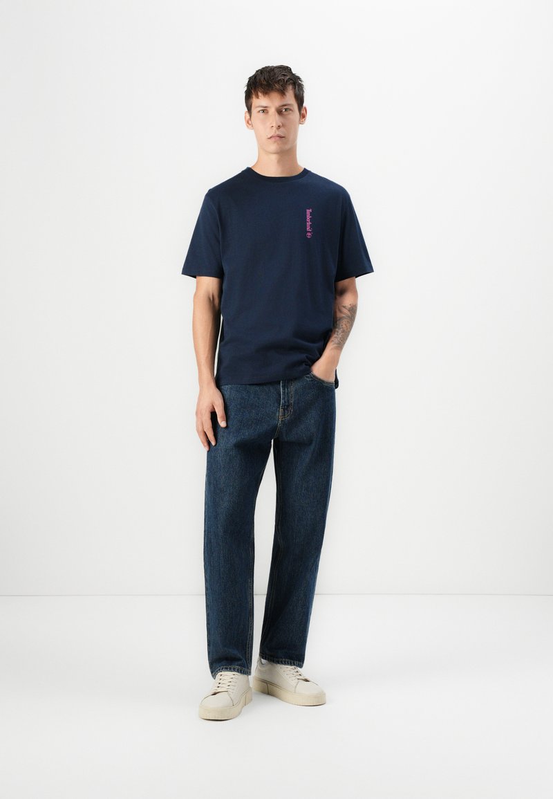 Młody mężczyzna stoi na tle białej ściany, ubrany w granatową koszulkę z grafiką, ciemnoniebieskie dżinsy i białe sneakersy, z tatuażem widocznym na ramieniu.