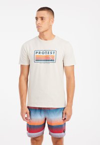 Crème t-shirt met de grafische tekst "PROTEST" en strepen boven "SUN SEA SURF." Kleurige zwembroek met horizontale gradientlijnen in oranje en blauw.