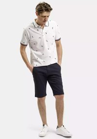 Biała koszulka polo z małymi czarnymi wzorami, w paski kołnierzyk; połączona z ciemnymi szortami i białymi trampkami. Gładka tkanina, luźny krój.