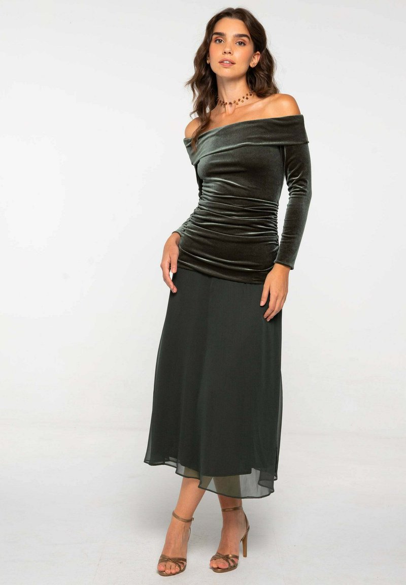 Robe décolletée en velours vert foncé avec des détails froncés ; jupe fluide en tissu transparent ; associée à des talons bruns à lanières et un collier simple.