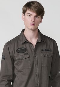 Bruine button-up shirt gemaakt van stof met een gladde textuur. Het heeft twee borstzakken en geborduurde patches op de borst en mouw.