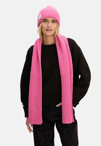 Różowa dziana czapka i pasujący do niej szalik z prążkowanej wełny. Model ma na sobie czarny sweter z okrągłym dekoltem i luźnymi rękawami.