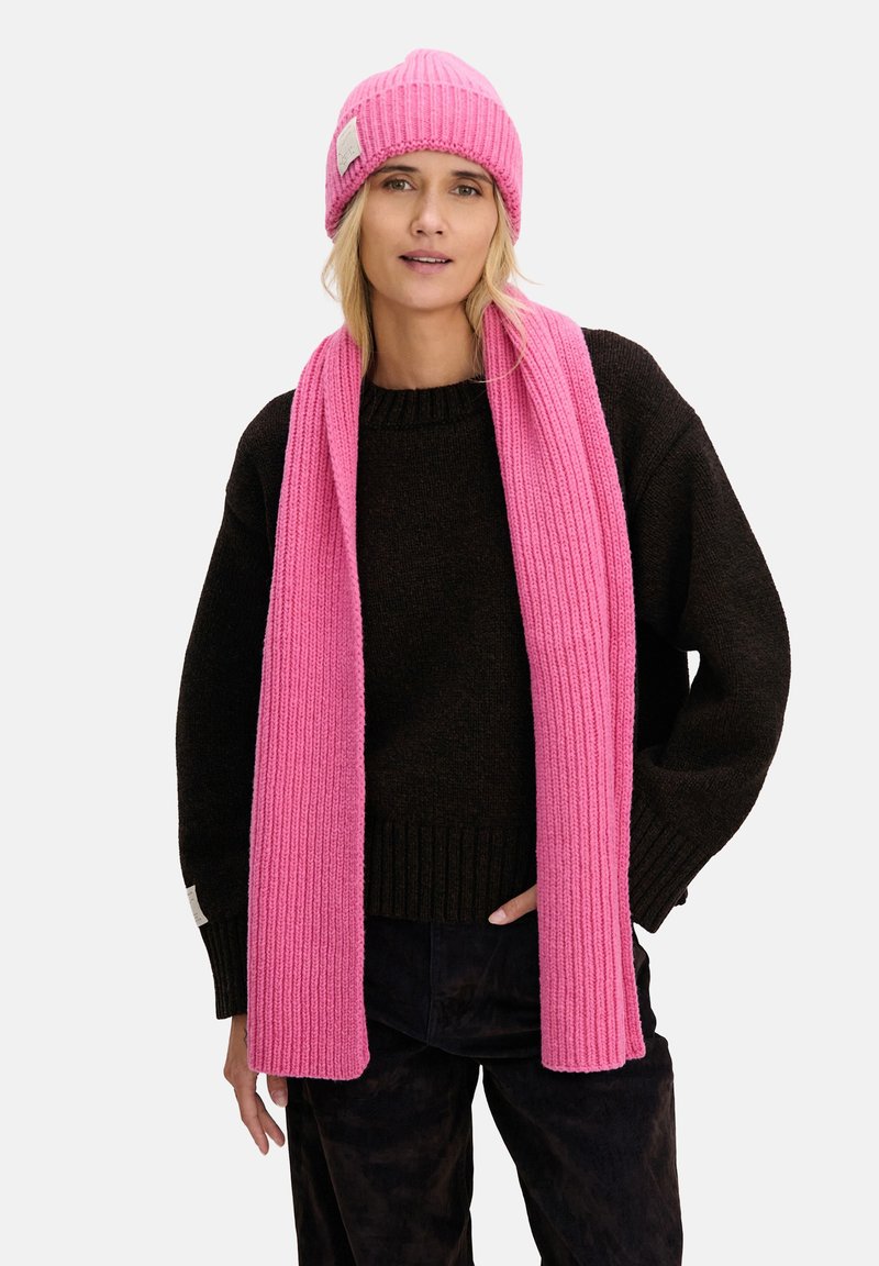 Różowa dziana czapka i pasujący do niej szalik z prążkowanej wełny. Model ma na sobie czarny sweter z okrągłym dekoltem i luźnymi rękawami.