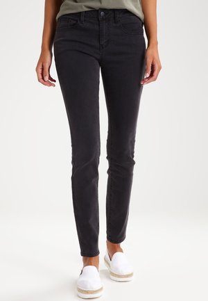 Person iført slanke sorte jeans og hvide slip-on sko, stående mod en ensfarvet lys baggrund.