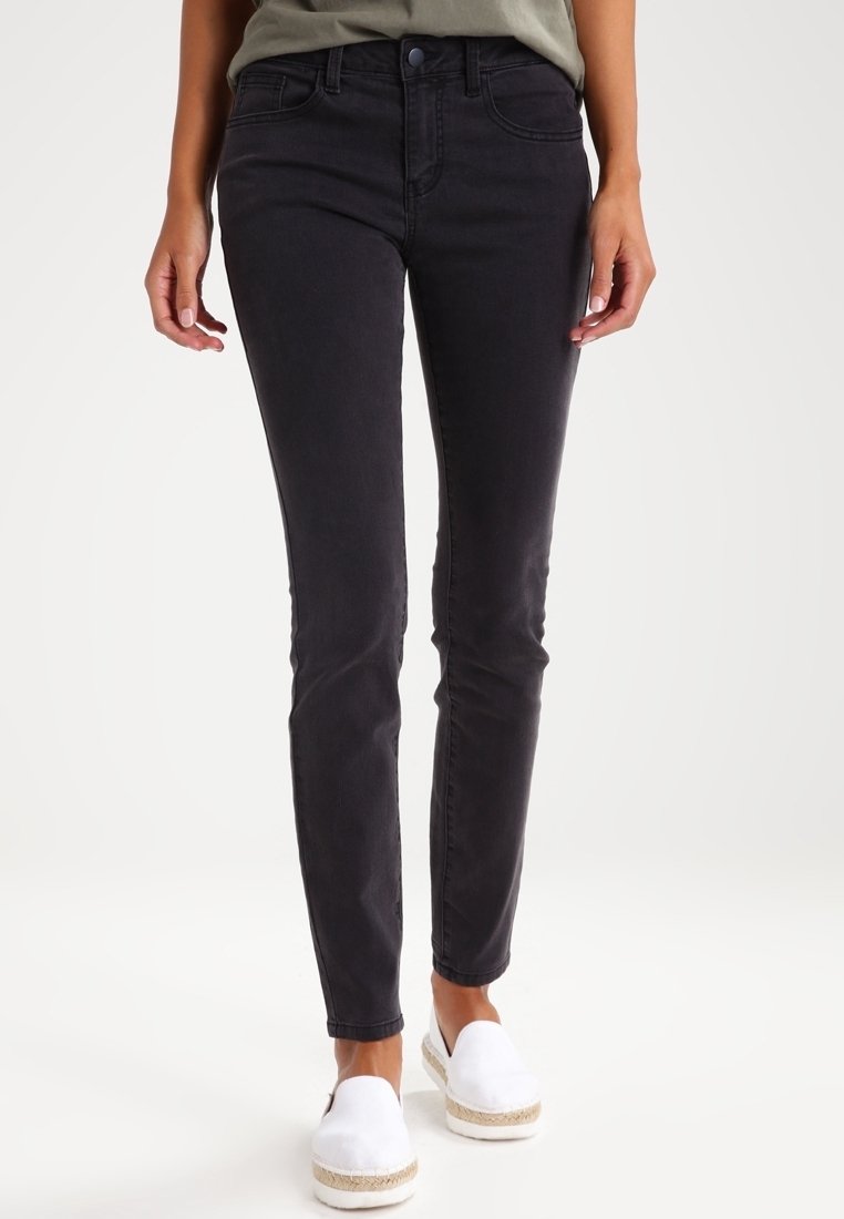 Personne portant un jean noir slim et des chaussures blanches sans lacets, debout devant un fond clair uni.