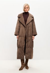 Cappotto lungo imbottito marrone con grande colletto, chiusura a bottoni e vita arricciata. Indossato sopra un top scuro con stivali con tacco a stampa leopardata.