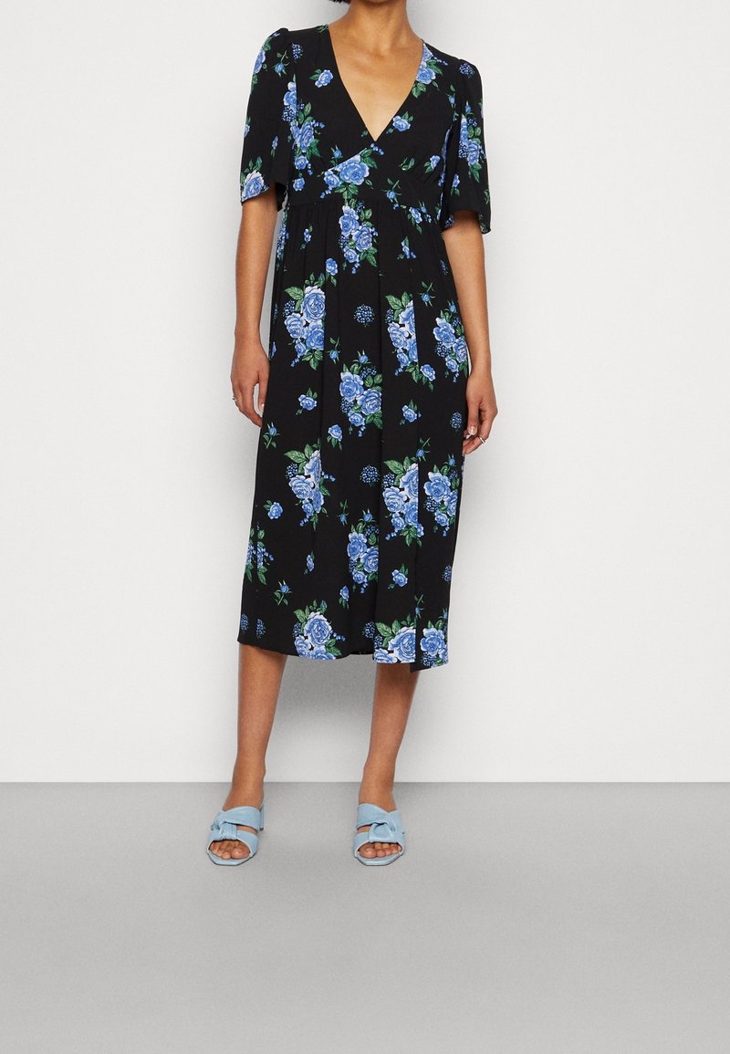 Robe midi noire à imprimé floral bleu, col en V, manches courtes, portée avec des sandales ouvertes à talon plat bleu clair.