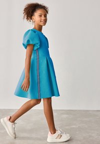 Helles blaues Kleid mit Puffärmeln, strukturiertem Muster und weißen Streifenakzenten. Kombiniert mit weißen Sneakers mit pinken und goldenen Details.