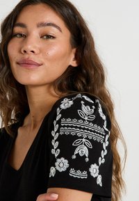 Camisa negra de cuello en V con bordado floral blanco en los hombros, que presenta patrones intrincados y detalles de costura texturizados.