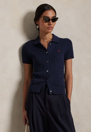 Kvinne iført marineblå kortermet kabelstrikket poloshorte med knapper, svarte vide bukser, kattøye-solbriller og hengende øredobber, stående mot en vegg.