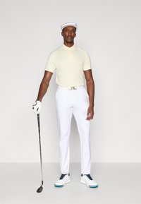 Polo shirt in lichtgeel, witte broek en witte schoenen met blauwe accenten. Houden van een golfclub, dragen van een witte pet en golfhandschoen.