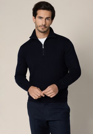 Mężczyzna w czarnym swetrze z długim rękawem i zamkiem przy szyi, noszący białą koszulę, stojący na tle jednolitego beżowego tła.