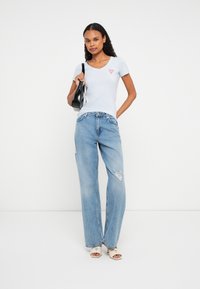 Kobieta z kręconymi włosami, ubraną w jasnoniebieską koszulkę z logo, podarte niebieskie dżinsy, białe sandały na obcasie i trzymającą czarną torbę na ramieniu.