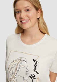 Lächelnde Frau mit blonden Haaren, die ein weißes T-Shirt trägt, das ein besticktes abstraktes Gesicht und den Text "Sei du selbst" zeigt.