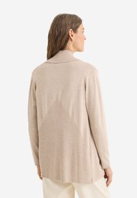 Cardigan beige con texture a coste e un elemento di design triangolare sul retro, con colletto a risvolto e maniche lunghe.