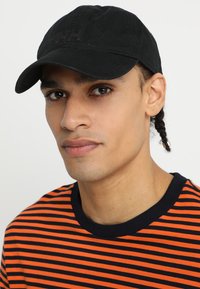 Gorra de béisbol negra con un tejido texturizado y detalle de logo, llevada con una camiseta a rayas naranja y navy, con un cuello redondeado.