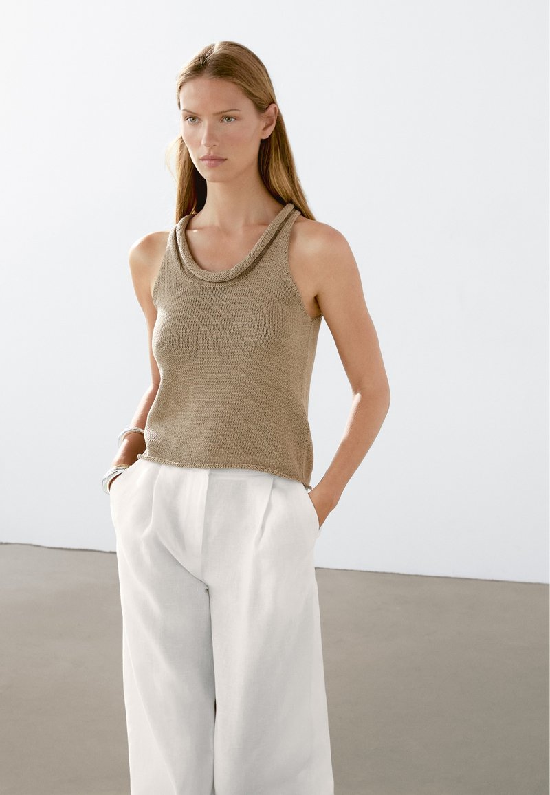 Beige gestricktes Tanktop mit einem schalenförmigen Ausschnitt, kombiniert mit weißen, weit geschnittenen Leinenhosen. Glatte Textur mit einer lockeren Passform.