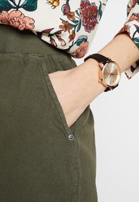Jupe vert olive avec une finition texturée, dotée d'une poche sur le côté avec un bouton. Une montre dorée avec un bracelet noir est portée au poignet.