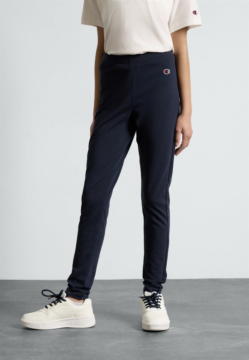 Navyfarbene enganliegende Jogginghosen mit elastischem Bund und kleinem Logo-Detail. Kombiniert mit weißen Sneakers, die einen glatten, dehnbaren Stoff zur Schau stellen.