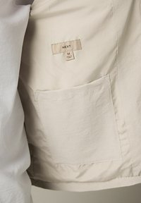 Beige jasvoering met een label waarop "NEXT" staat, maat M, inclusief een rechthoekig zakje in de stof genaaid. Zachte, gladde textuur.