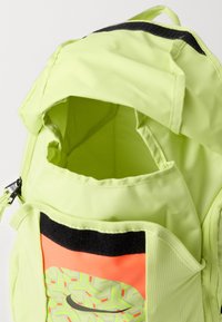 Rucsac verde-însorit cu galben deschis, cu compartiment principal deschis, bandă neagră din Velcro și buzunar frontal cu un model portocaliu geometric și logo Nike.