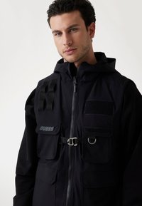 La chaqueta negra con capucha presenta cierre de cremallera, múltiples bolsillos frontales, correa ajustable y parches de tela. La textura parece suave y mate.