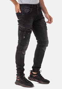Jeans noirs ajustés avec plusieurs poches, fermetures éclair et détails de couture. Fabriqués en denim, avec une finition texturée et des poignets élastiques.