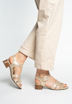 Feet wearing beige strappy block-heeled sandals with a buckle, paired with rolled-up light beige corduroy pants against a neutral background.