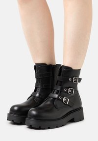 Černé kožené kotníkové boty s kulatou špičkou, vybavené třemi nastavitelnými pásky se stříbrnými přezkami a bočním zipem. Robustní gumová podrážka.