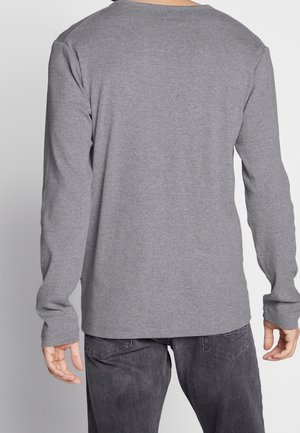 Personne portant un t-shirt gris uni à manches longues et un jean gris foncé, vue de dos avec les bras détendus le long du corps.