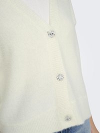 Cream cardigan with a v-neck, ribbed texture, and three decorative buttons featuring silver accents and a clear center gem.
