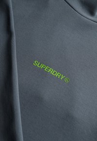 Sudadera gris con una textura suave, que presenta un logotipo "SUPERDRY" en brillante color verde en el frontal. Diseño sencillo con cuello redondo.