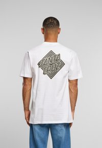 Balta kokvilnas T-kreklu ar lielu melnu ģeometrisku logo "SANTA CRUZ", kas izdrukāts aizmugurē. Īsas piedurknes, brīvs piegriezums.
