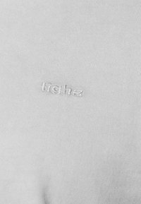 Hellgrauer Baumwollstoff mit subtilen vertikalen Rippungen. Enthält gesticktes "tigha"-Logo in passendem Faden.