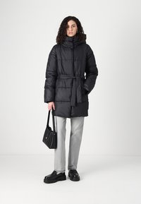 Črna puffer jakna z visokim ovratnikom in pasom; vključuje prešito zasnovo, kombinirano s sivo jeans hlačami, črno torbico in platform čevlji.