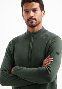 Groene gebreide pullover met een halve rits kraag, geribbelde manchetten en een logo patch op de mouw. Gladde textuur en aansluitend ontwerp.